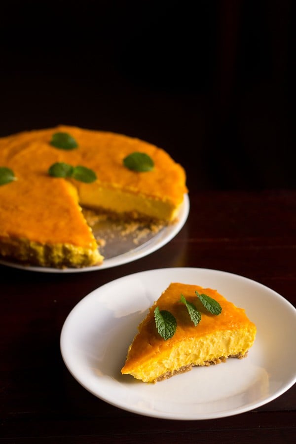 one slice of eggless mango cheesecake on a white plate with the rest of the cheesecake in the background.