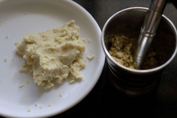 crumbled khoya on a plate next to a grinder bowl full of ground nuts