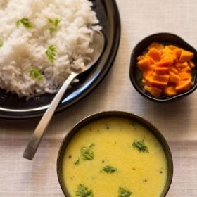 maharashtrian kadhi garnished with coriander leaves and served in a black bowl with some poured over steamed rice kept on a plate with a spoon on the upper left side and a small bowl of carrots kept on the upper right side.