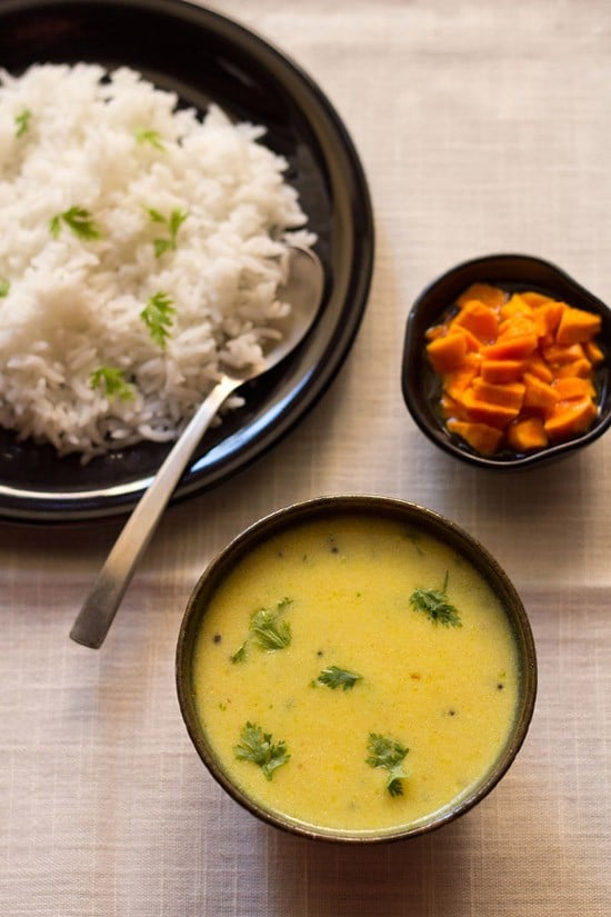 maharashtrian kadhi garnished with coriander leaves and served in a black bowl with some poured over steamed rice kept on a plate with a spoon on the upper left side and a small bowl of carrots kept on the upper right side.