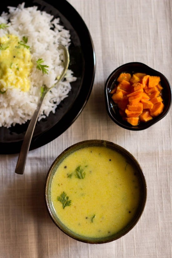 The Maharashtrian kadhi is garnished with coriander leaves and served in a black bowl topped with some rice and placed on a plate with a spoon on the upper left and a small bowl of carrots on the upper right. 