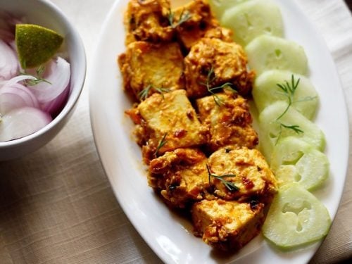 malai paneer served on a white platter with cucumber slices kept on left side on the platter, a bowl of onion and lemon.