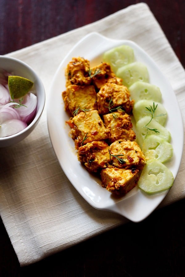 top shot of malai paneer served on a white platter with cucumber slices kept on right side on the platter, a bowl of onion and lemons. 