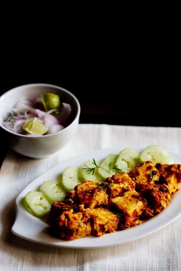 malai paneer served on a white platter with cucumber slices kept on left side on the platter, a bowl of onion and lemons.