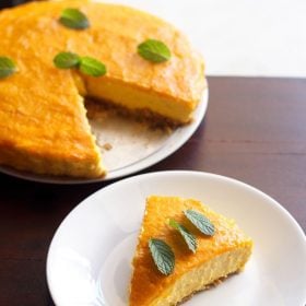 one slice of eggless mango cheesecake on a white plate with the rest of the cheesecake in the background.