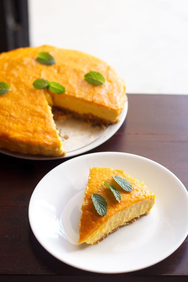 one slice of eggless mango cheesecake on a white plate with the rest of the cheesecake in the background.