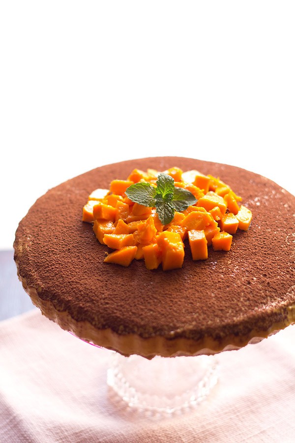 no-bake eggless mango cheese cake on a cake stand, topped with cocoa powder, fresh mango cubes, and a sprig of mint.