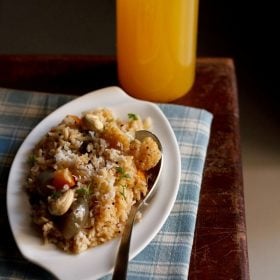 masale bhat garnished with grated coconut, roasted cashews, chopped coriander leaves and served on a white plate with a spoon on the right side and a glass of juice kept in the background.