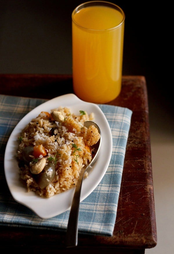 masale bhat garnished with grated coconut, roasted cashews, chopped coriander leaves and served on a white plate with a spoon on the right side and a glass of juice kept in the background.