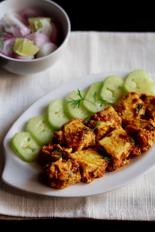 malai paneer served on a white platter with cucumber slices kept on left side on the platter, a bowl of onion and lemons. 