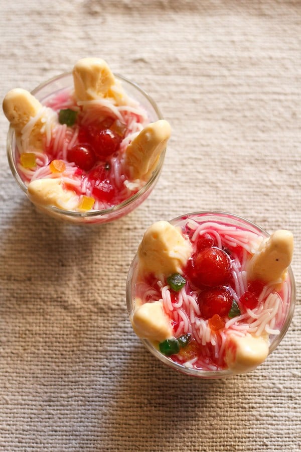 mango kulfi falooda topped with rose syrup, red cherries, tutti frutti in two bowls on a cream jute cloth