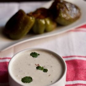 onion raita in a white bowl garnished with mint leaves with a background of stuffed bell peppers in the background