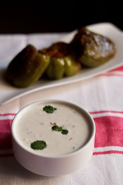 onion raita in a white bowl garnished with mint leaves with a background of stuffed bell peppers in the background