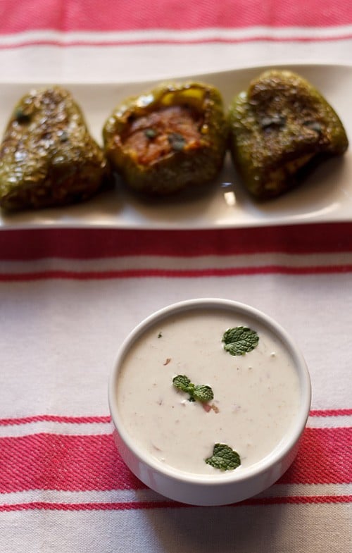 top shot of onion raita served in a bowl, garnished with mint leaves with a white tray of stuffed bell peppers placed at the top