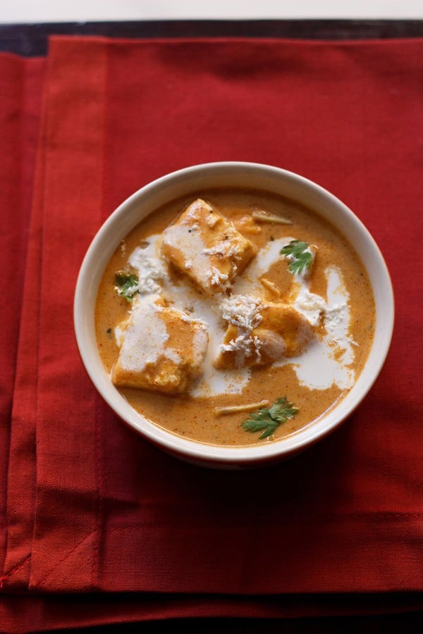 overhead shot of paneer makhanwala topped with cream, grated paneer, cilantro in an off white bowl on a red cotton napkin