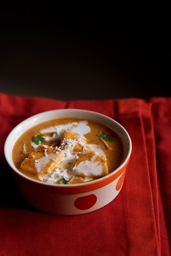 paneer makhanwala topped with cream, grated paneer, coriander leaves in an orange dotted bowl on a red cotton napkin.