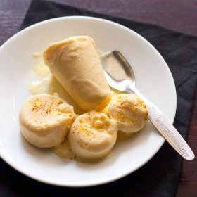 mango kulfi sliced and whole on a white plate with a text layover with a steel spoon