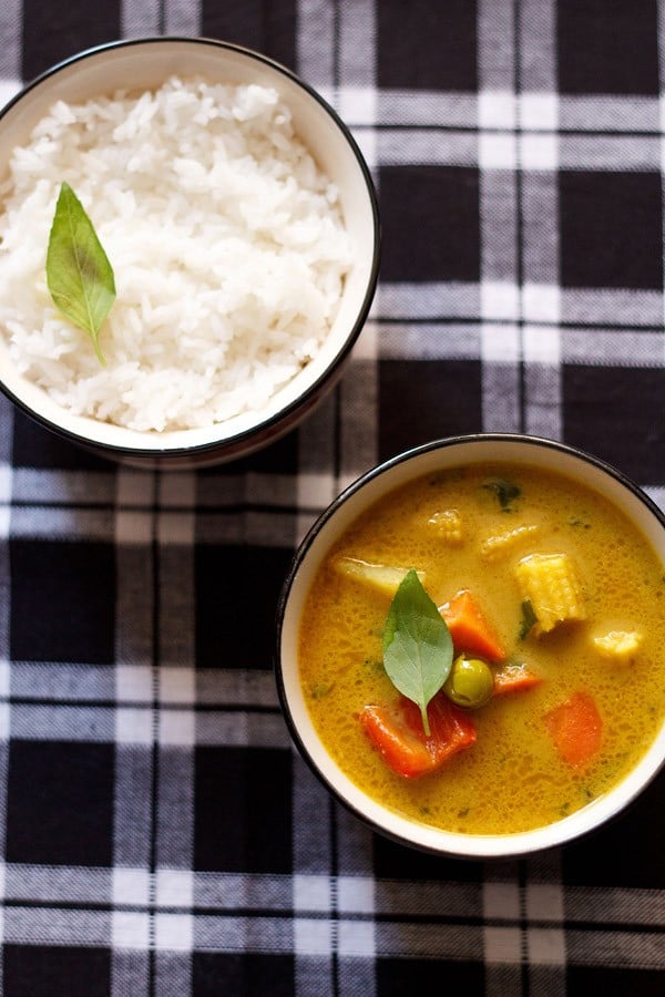 thai yellow curry garnished with a basil leaf and served in a black rimmed ceramic bowl with a bowl of steamed rice kept on the top left side. 