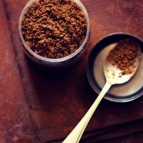 goda masala in a jar with spoon filled with the spice blend on left side.