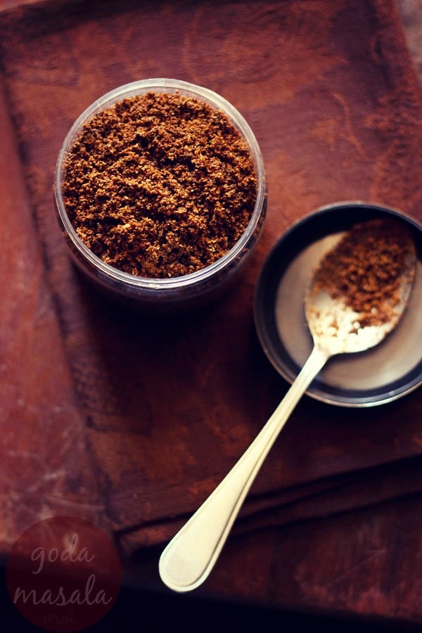 goda masala in a jar with spoon filled with the spice blend on left side.