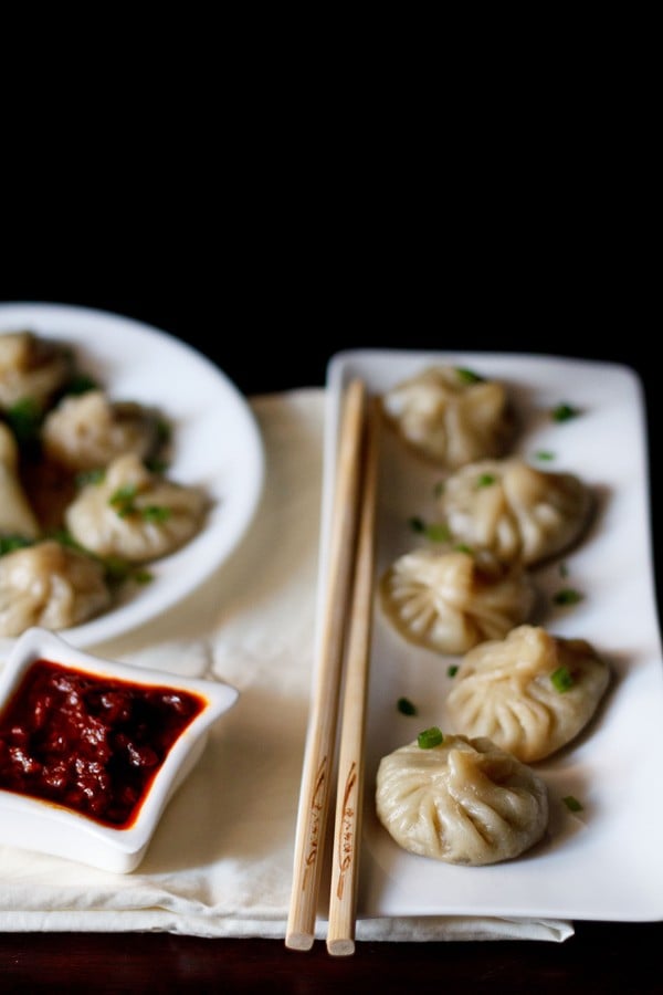 Veg momos arranged on a white rectangular long tray with a bamboo chopsticks by the side and a small square bowl of schezwan sauce at the side