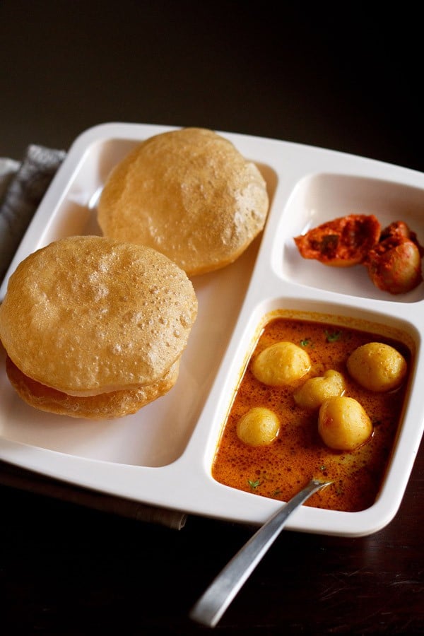 dum aloo banarasi served in a plate with pooris, lemon pickle and a spoon dipped in it. 