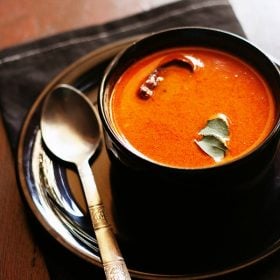 tomato curry garnished with curry leaves and served in a black bowl kept on a black plate with a spoon kept on left side of the bowl.