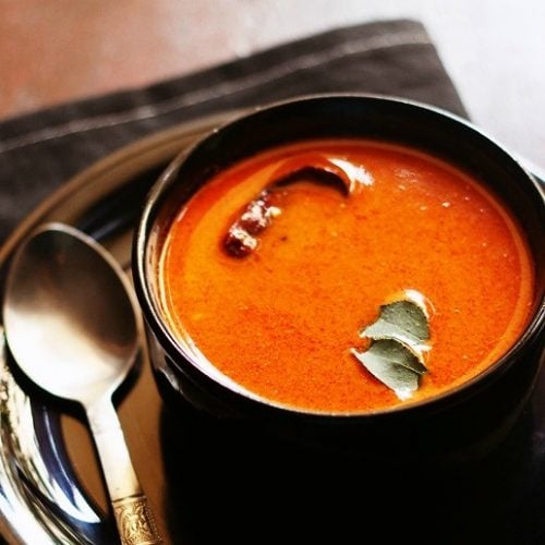 tomato curry garnished with curry leaves and served in a black bowl kept on a black plate with a spoon kept on left side of the bowl.