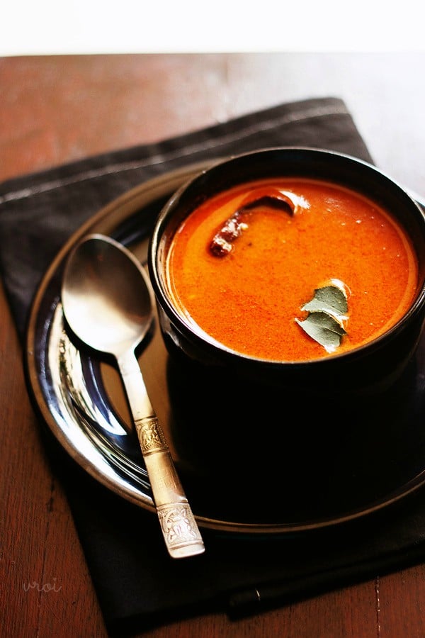 tomato curry garnished with curry leaves and served in a black bowl kept on a black plate with a spoon kept on left side of the bowl.