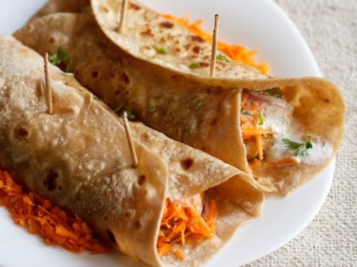 two rolled veg frankie on a bed of grated carrots on a square shaped white plate on a burlap
