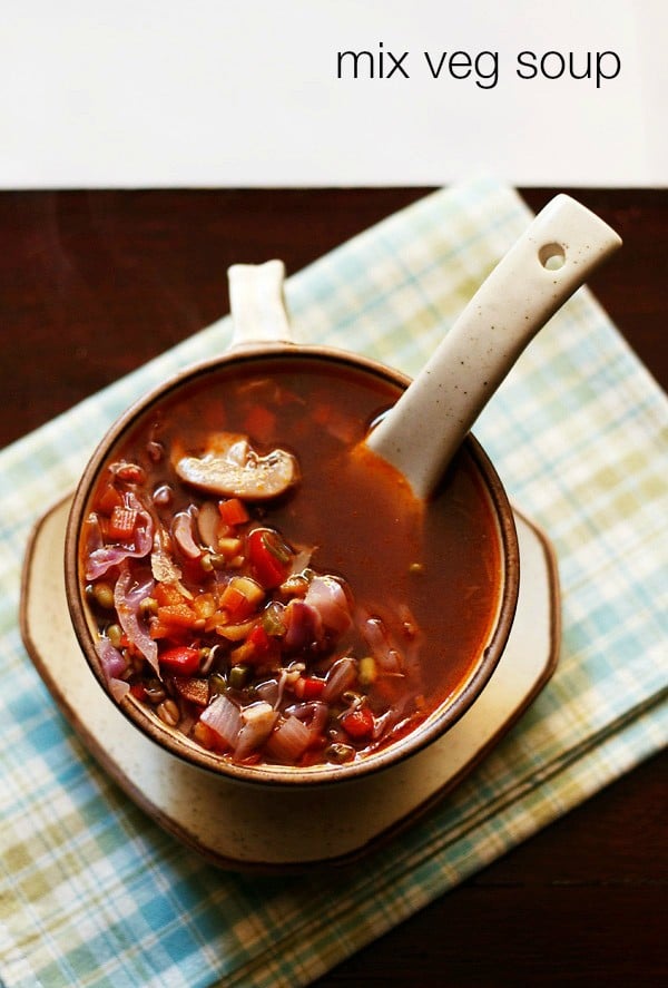 mix vegetable soup in a cream colored ceramic mug with a ceramic spoon inside the bowl on a checkered green, white, blue napkin 