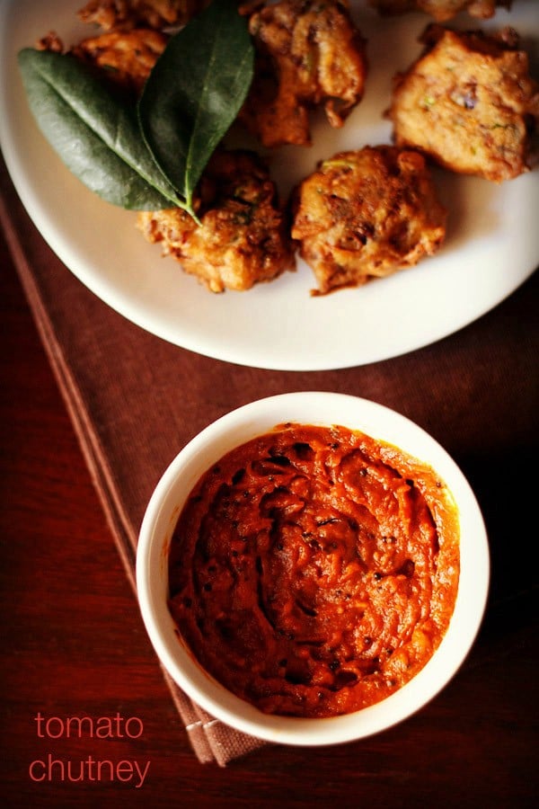 tomato chutney served in a small white bowl with a platter of pakoda kept on the top side and text layover.