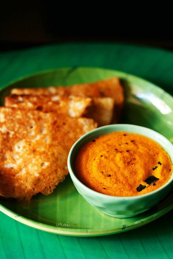 red coconut chutney served in a green colored bowl placed on a green colored plate with folded dosas kept on the left side. 