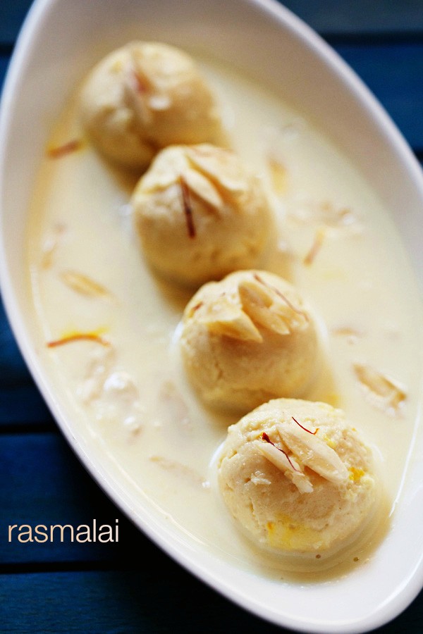 4 balls of rasmalai in an oblong white serving dish covered with thickened milk and garnished with sliced almonds and saffron threads