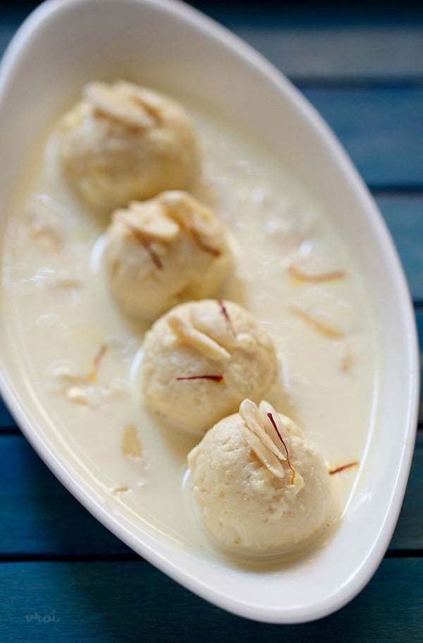 4 balls of rasmalai in an oblong white serving dish covered with thickened milk and garnished with sliced almonds and saffron threads
