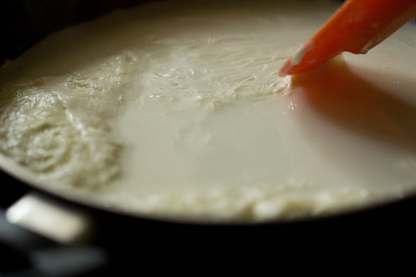 simmering milk for rasmalai recipe has the cream and milk solids floating on top and being pushed to the side with a spatula
