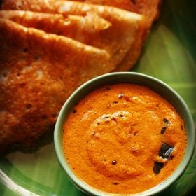 red coconut chutney served in a green colored bowl placed on a green colored plate with folded dosas kept on the upper left side and text layovers.