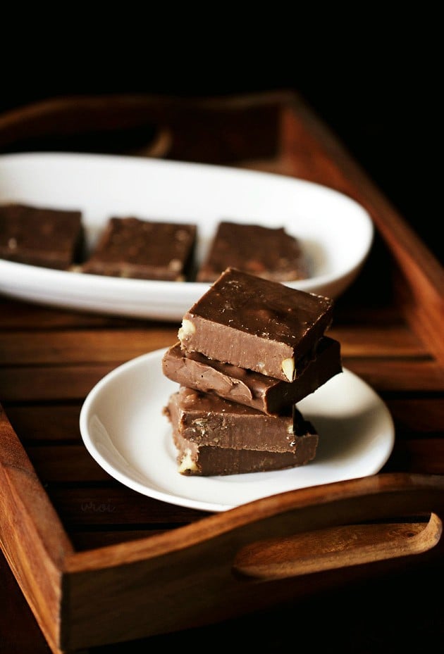 Chocolate fudge squares stacked on top of each other placed on a white plate on a wooden tray. 