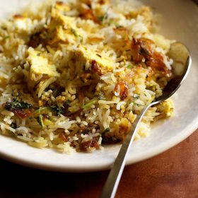 paneer biryani served in a cream shallow plate with a steel spoon by the side on a brown wooden board