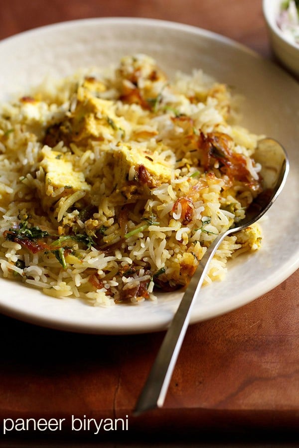 paneer biryani served in a cream shallow plate with a steel spoon by the side on a brown wooden board