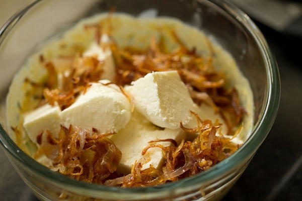 paneer cubes and caramelized onions added to the whisked curd in the glass bowl