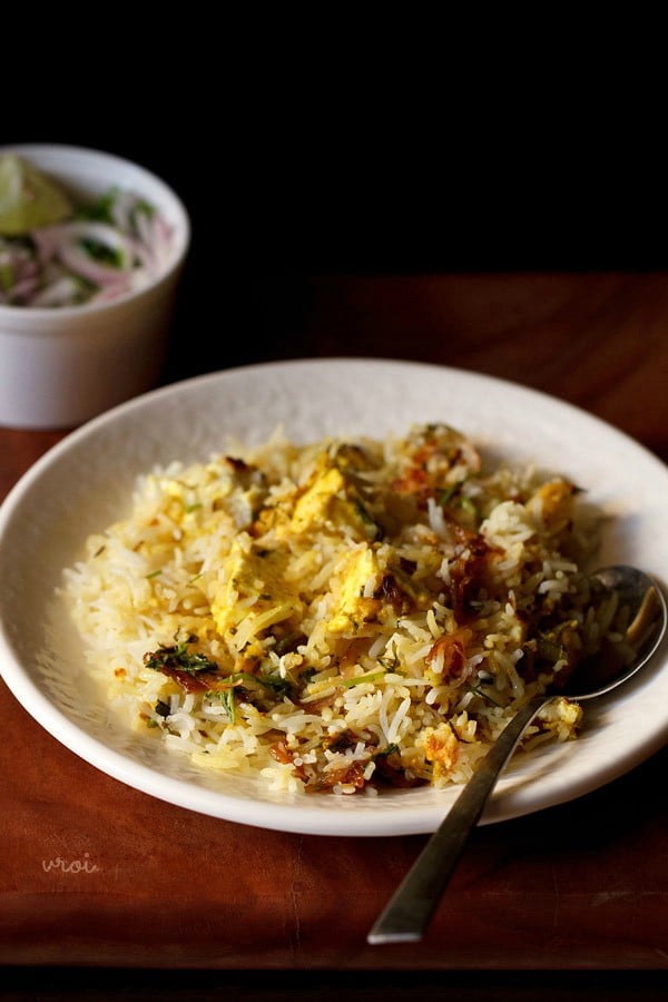 paneer biryani served in a cream shallow plate with a steel spoon by the side on a brown wooden board