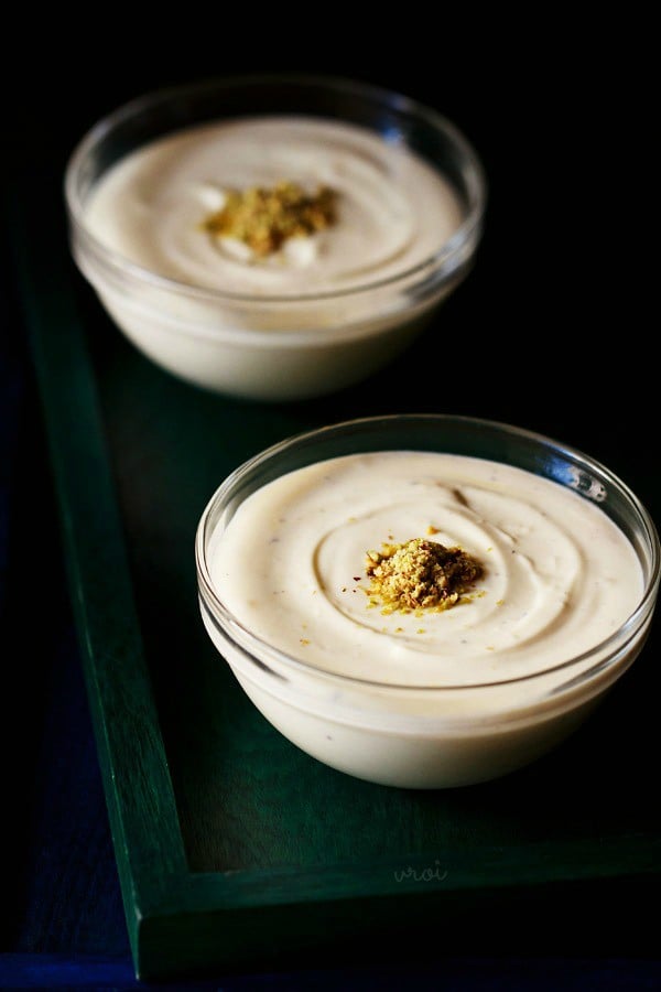 two small glass bowls of kesar elaichi shrikhand topped with chopped pistachios on a dark green serving tray