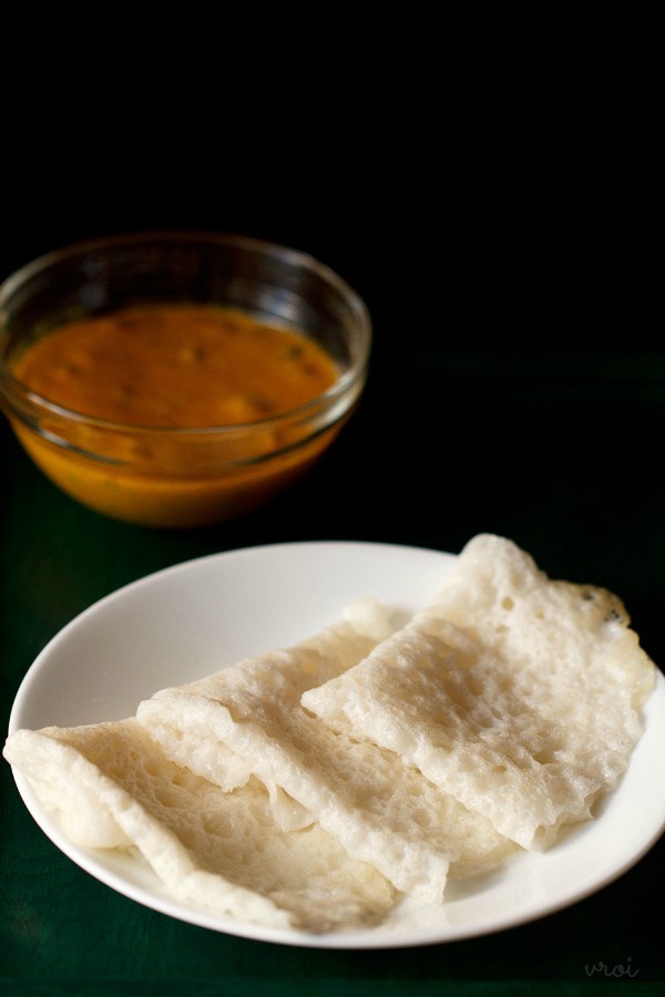 three folded neer dosas on a white plate with a bowl of coconut chutney in the background