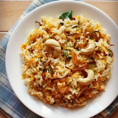 carrot rice garnished with cashews, fried onions and raisins served in a white plate on a checkered white and blue napkin