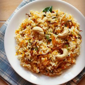 carrot rice garnished with cashews, fried onions and raisins served in a white plate on a checkered white and blue napkin.