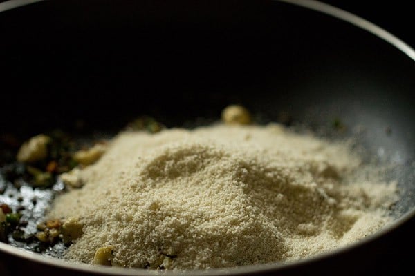 Rava added on top of ginger, nut, and seasoning mixture sautéing in black skillet.