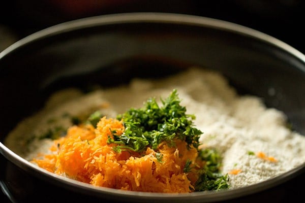 Grated carrots and coriander leaves added to oats idli in black bowl.