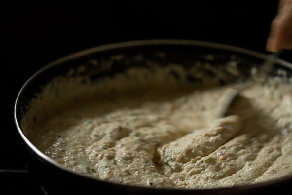 Person using spoon to stir oats idli batter in black skillet.