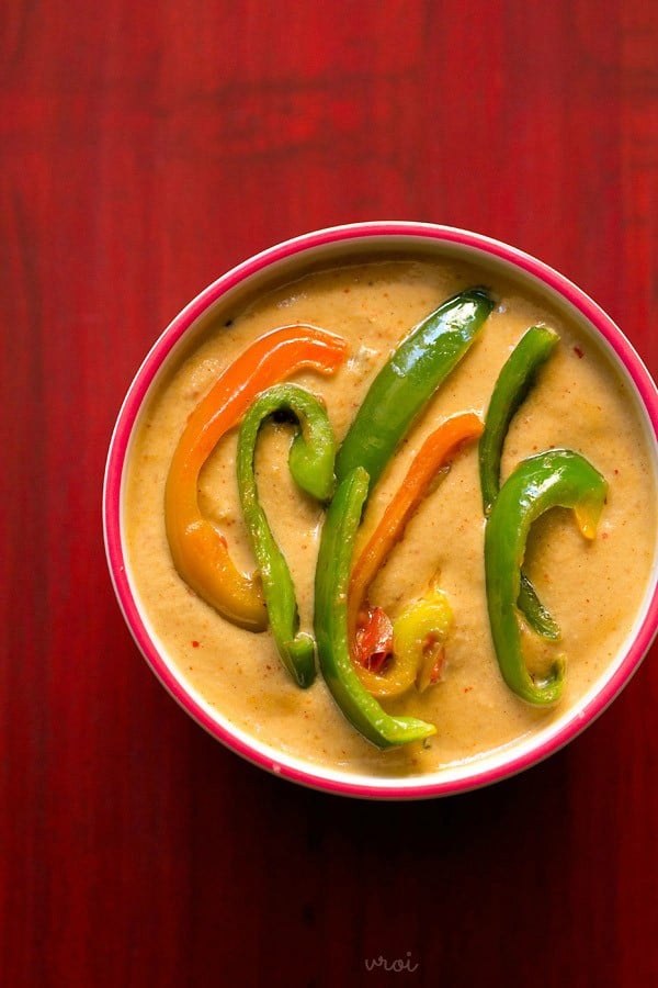 capsicum masala gravy with sliced of red and green capsicum in a dark pink rimmed bowl on a red wooden board